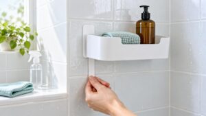 Hand pulling a white adhesive release strip beneath a white shower caddy mounted on wet tiled bathroom walls, holding an amber pump bottle and folded mint-green cloth.